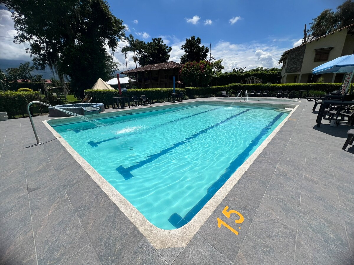 An outdoor swimming pool is displayed, featuring clear blue water and marked depth indicators. Surrounding areas include lounge chairs with dark frames and tables set beneath umbrellas. Lush greenery borders the pool area, contributing to a serene atmosphere.