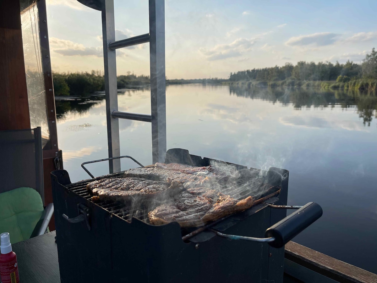A charcoal grill is positioned on the deck of a houseboat, with meat sizzling and smoke gently rising. The serene waters of the Peene reflect a soft sunset, surrounded by lush greenery. The scene captures a peaceful outdoor cooking experience.