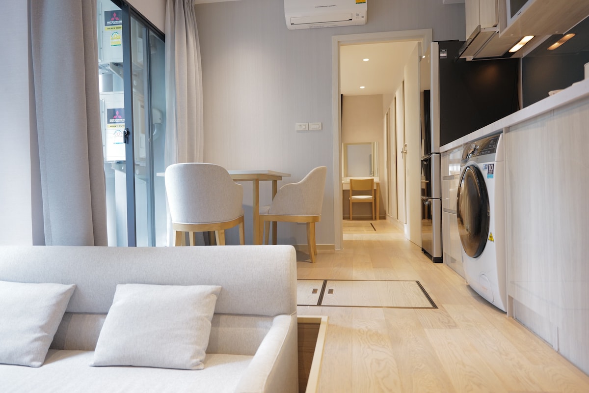 A bright living area features a light-colored sofa with decorative cushions. In the background, a small dining table is set for two, with additional seating nearby. A modern kitchen area is visible, equipped with a washing machine and sleek cabinetry.
