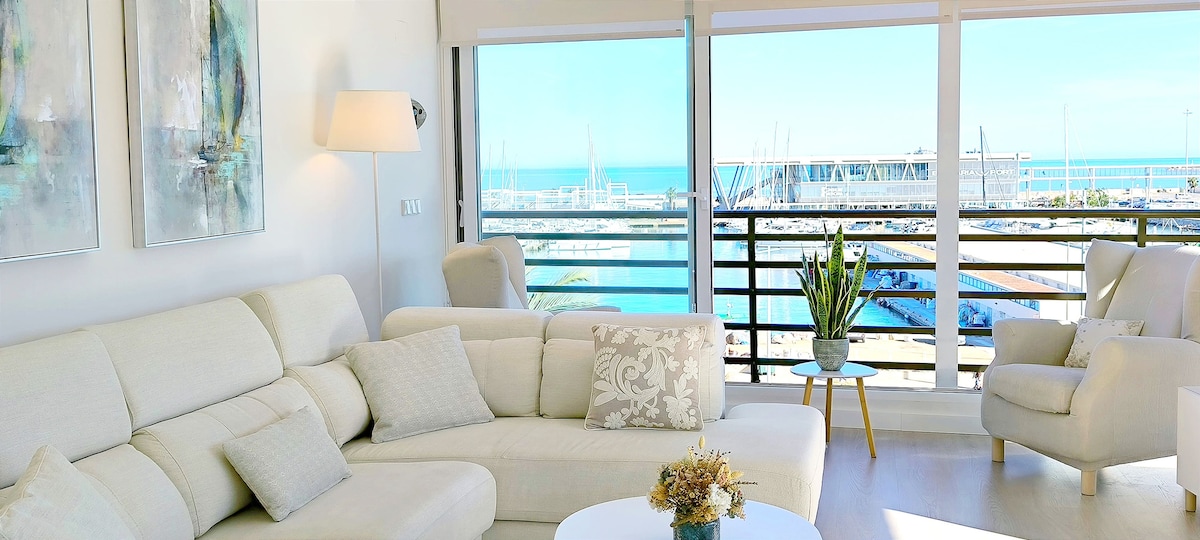 A spacious living area features large windows providing a clear view of the harbor. A comfortable white sectional sofa is positioned to allow for relaxation while enjoying the scenery. A round coffee table sits at the center, complemented by decorative elements and a plant nearby.