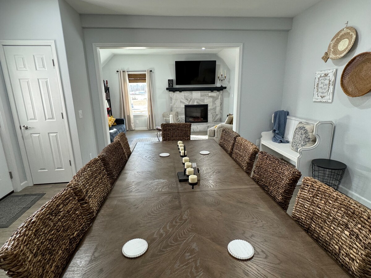A long farmhouse table is surrounded by woven chairs, set for communal gatherings. The room features a stone fireplace in the background, with light streaming through large windows, inviting a sense of warmth and spaciousness.