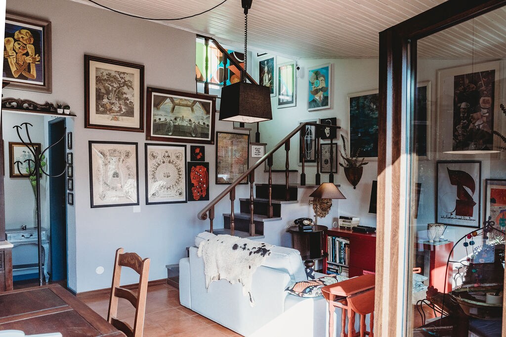 A light-filled living area features an eclectic display of framed artwork on the walls. A staircase leads to the upper floor, while a cozy sofa and various decorative items are arranged around a wooden table, creating an inviting space for relaxation.