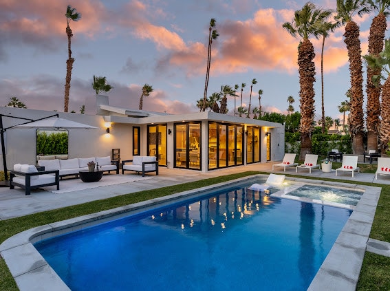 A modern home is framed by palm trees and set against a colorful sky. The heated pool shimmers in blue, with an attached spa nearby. Outdoor seating areas provide space for relaxation. Expansive glass doors reveal the interior, enhancing the connection between indoor and outdoor living.