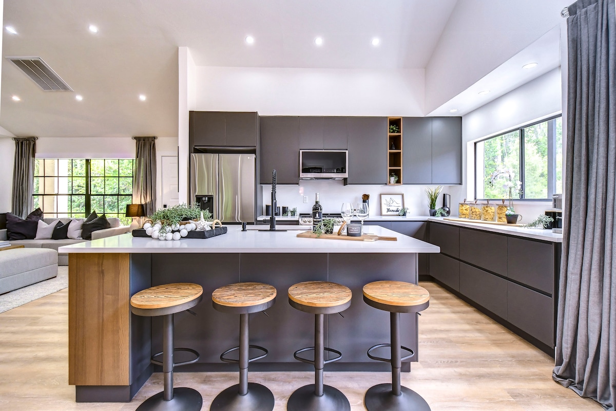 A modern kitchen is equipped with sleek cabinetry and spacious countertops. High-backed stools are arranged around a large island, while appliances and decorative items are neatly organized. Natural light floods in through windows, enhancing the inviting atmosphere.