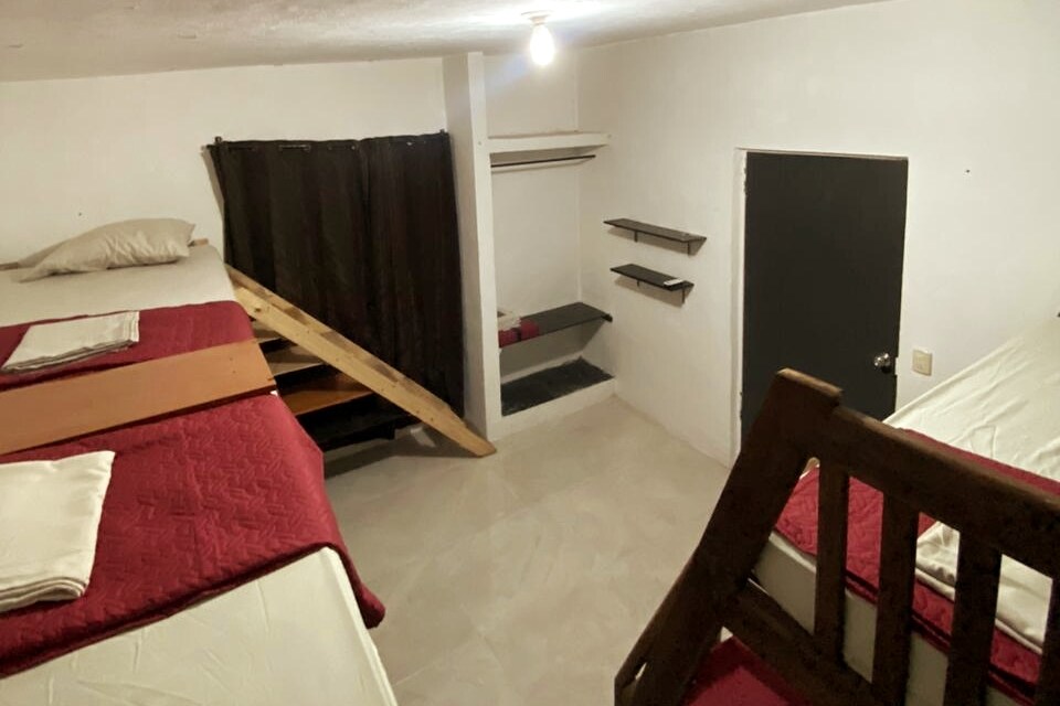 The sleeping area features multiple beds with red bedding spread across a spacious room. A wooden ladder leads to a lofted area, while shelves for storage are mounted on the wall. Soft lighting illuminates the space, enhancing visibility.