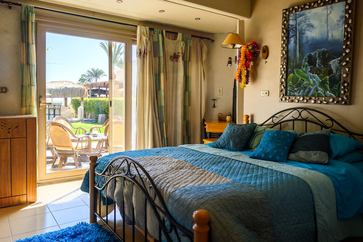 A cozy bedroom features a large bed with layered blankets, complemented by decorative pillows. A sliding glass door leads to the outdoor space, revealing a view of tropical plants and sitting areas. Natural light fills the room, enhancing its inviting and serene atmosphere.