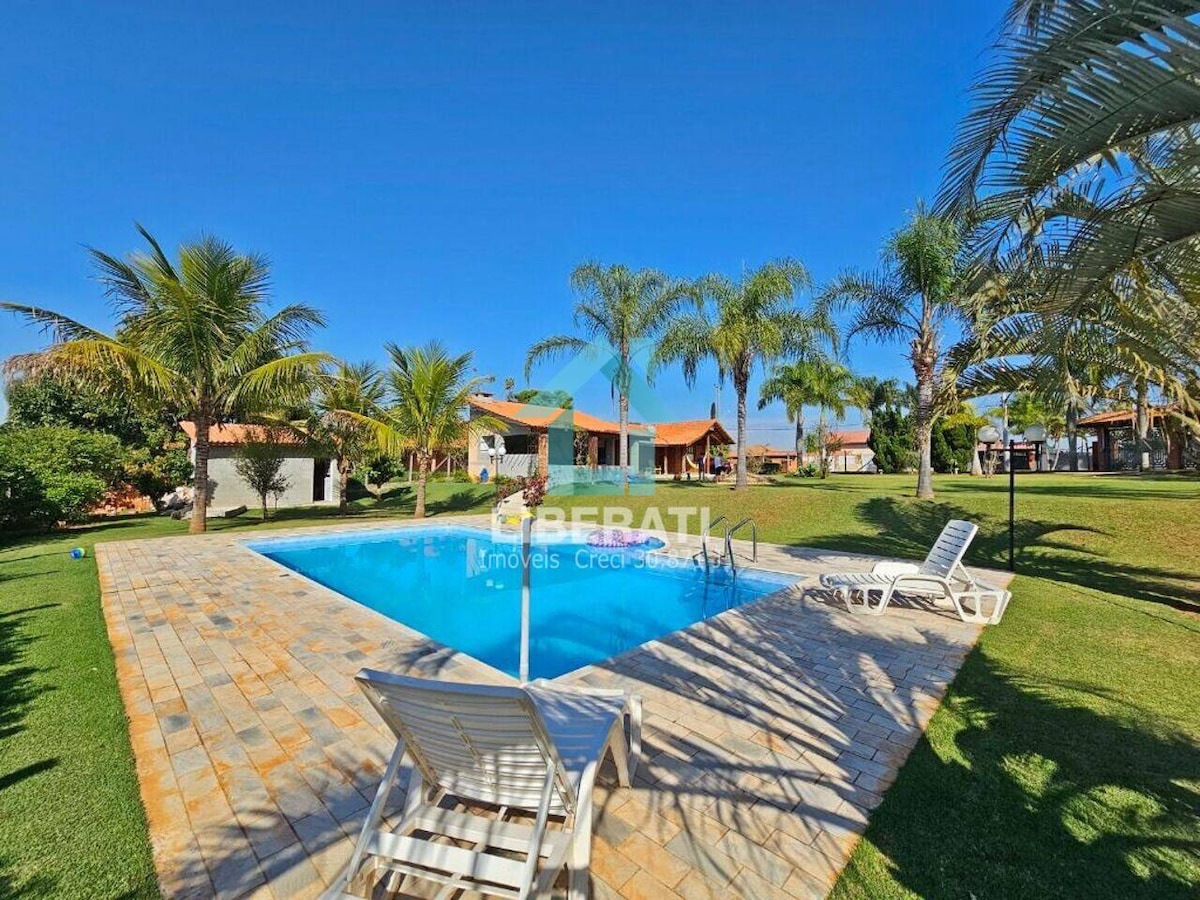 A serene outdoor area features a clear swimming pool surrounded by palm trees and well-maintained green lawns. Lounge chairs are positioned near the pool, offering a place to relax in the sun. Sunlight illuminates the scene, enhancing the inviting atmosphere.