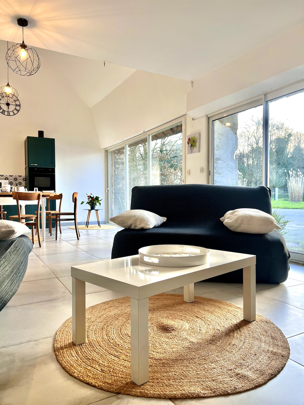 A bright living area is presented, featuring a dark sofa with decorative cushions. A round woven rug is positioned beneath a white coffee table. Large glass doors allow natural light to fill the space, offering a view of the green outdoor surroundings.