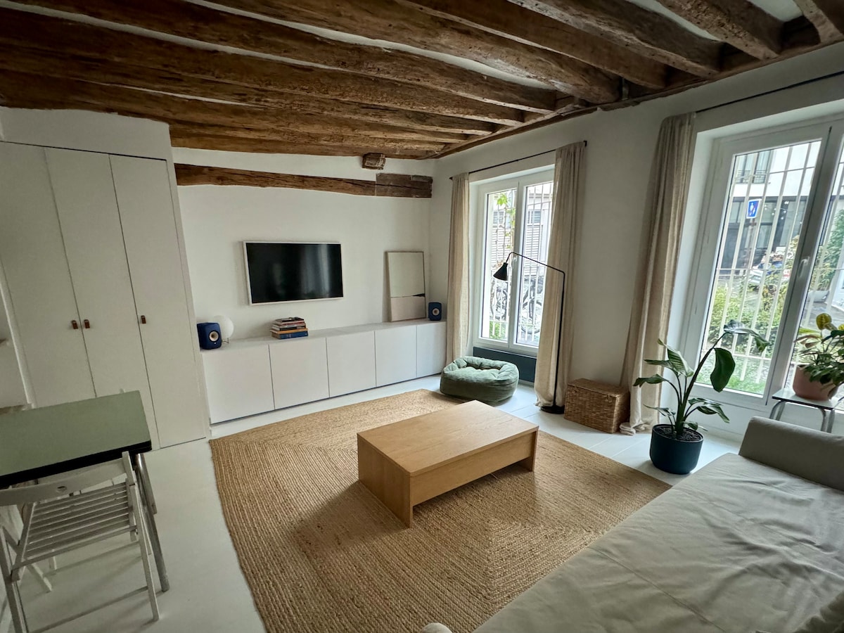 A spacious living area features wooden beams on the ceiling and large windows allowing natural light. A light-colored sofa faces a minimalist entertainment unit housing a television. A round green cushion lies on a textured rug, complemented by potted plants adding a touch of greenery.