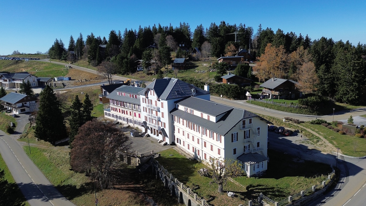 An aerial view of a multi-story building surrounded by a natural landscape, featuring a mix of greenery and residential structures. The building presents a symmetrical façade with prominent balconies, and a well-maintained parking area is visible in the foreground.