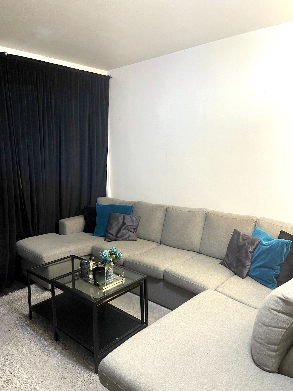 A light-flooded living area features a spacious gray sectional sofa adorned with cushions in varying shades of blue and black. A sleek glass coffee table sits in front, accented with a small decorative arrangement, and a dark curtain provides a contrast along one wall.
