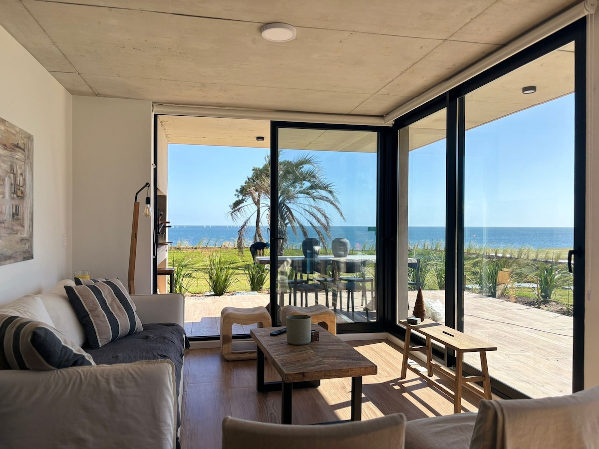 A bright living area features large glass doors that open to a patio with sea views. A light-colored sofa arranged with striped cushions is accompanied by a wooden coffee table. The space is complemented by natural light streaming in from the outdoor area.