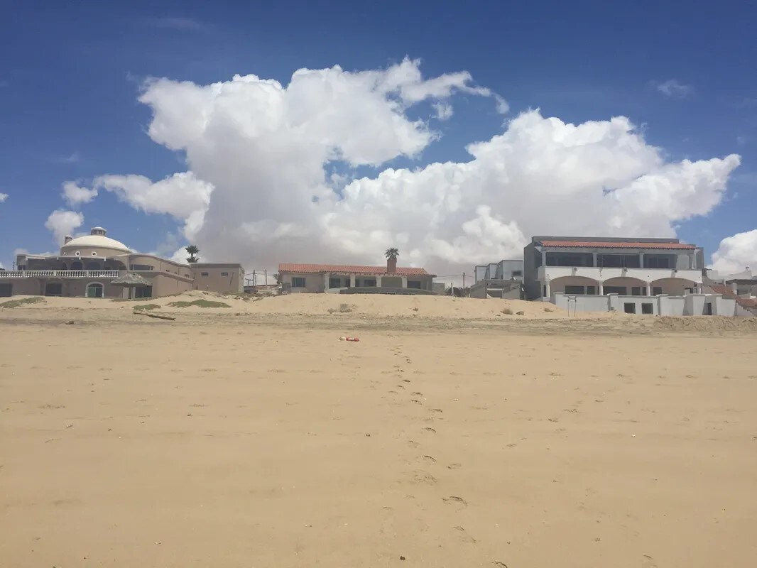 A shoreline view showcases several beach houses set against a backdrop of fluffy white clouds and a clear blue sky. The sandy beach stretches in the foreground, with footprints leading towards the homes, emphasizing the inviting atmosphere of this coastal location.