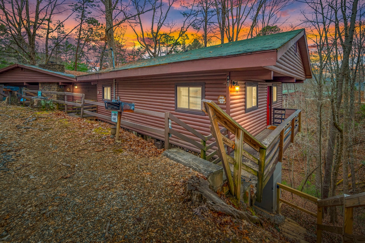 The duplex cabin features a rustic exterior with wood siding, blending seamlessly into the surrounding nature. A series of wooden steps lead up to the entrance, framed by trees, with a sunset sky casting warm colors across the scene.