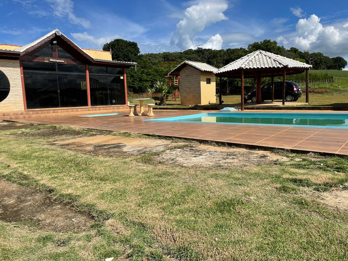 A spacious outdoor area features a rectangular pool surrounded by a tiled patio and green grass. Two nearby structures include a wooden gazebo with a thatched roof and a stone building. Blue skies and scattered clouds create a bright atmosphere.
