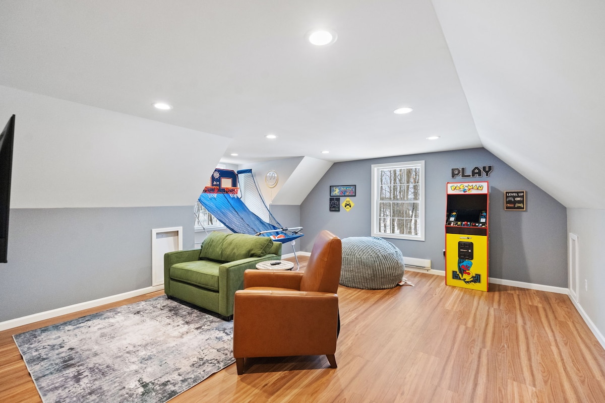 An inviting game room is showcased with light wood flooring and ample natural light. A green sofa, a brown armchair, and a large bean bag provide comfortable seating. Decor includes playful wall art, and a gaming area features a Pac-Man arcade machine against one wall.