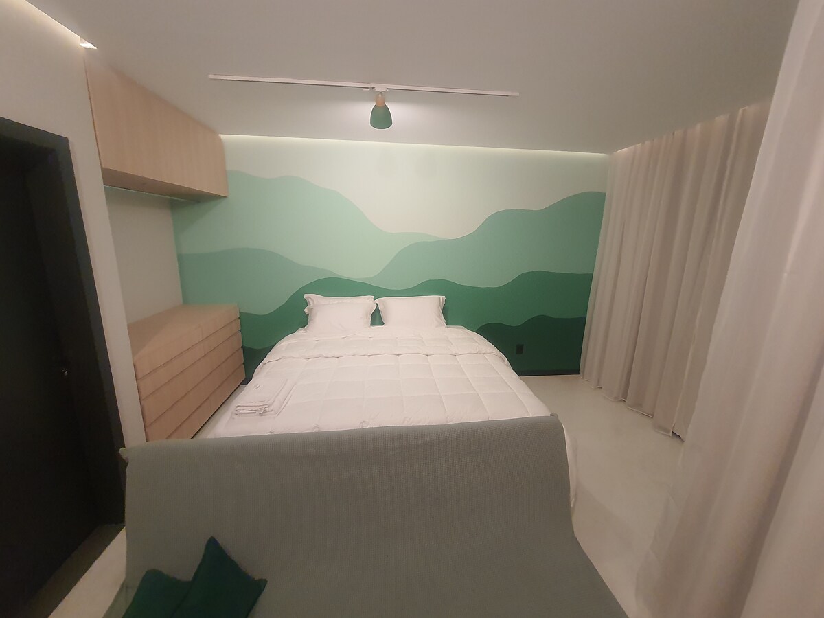 A calming bedroom features a spacious bed with crisp white linens against a soft green wall mural. Natural light filters through sheer curtains, complementing light wood cabinetry that provides storage. The room's minimalist design promotes a serene ambiance.