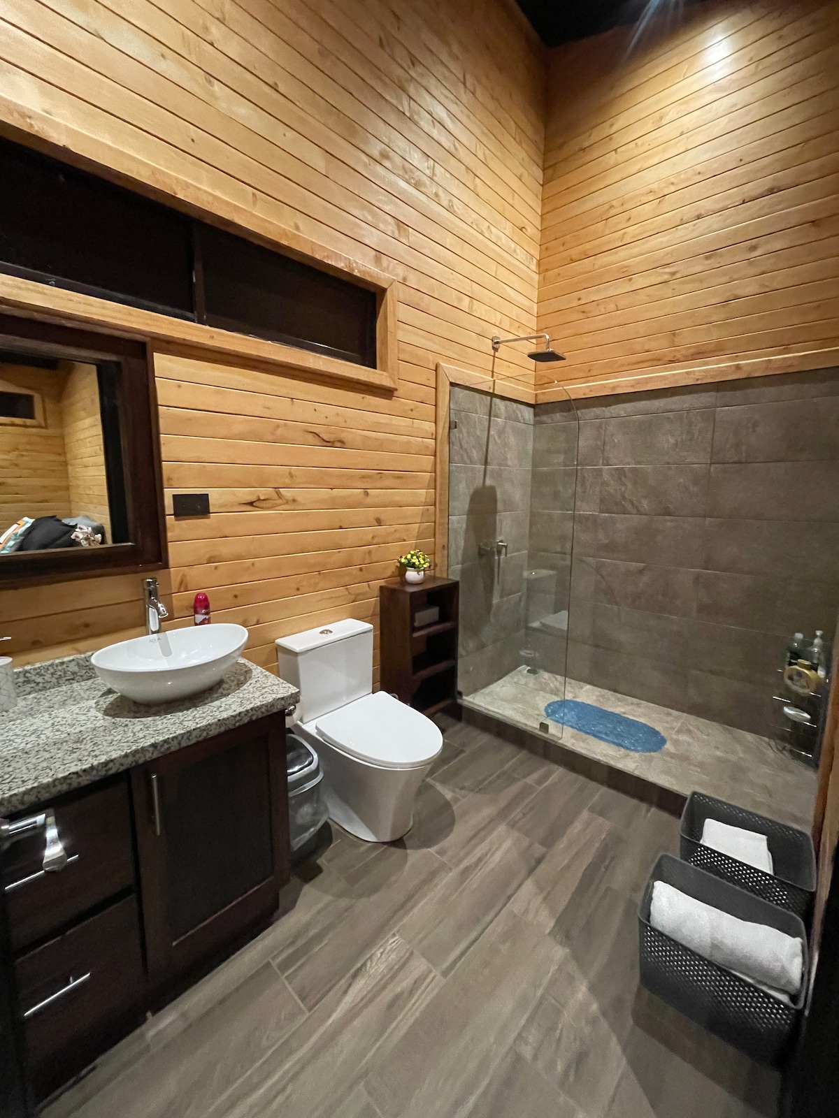The bathroom features warm wooden paneling and modern fixtures. A spacious shower with glass walls and a slate tile floor is highlighted. A sleek sink rests on a granite countertop, complemented by storage shelves and fresh towels neatly arranged nearby.