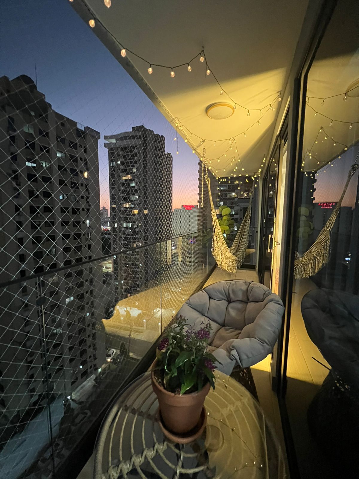 A balcony space features decorative string lights and a potted plant. A comfortable chair invites relaxation, while glass railings provide a clear view of the city skyline at dusk, showcasing the blend of urban architecture and the fading twilight sky.