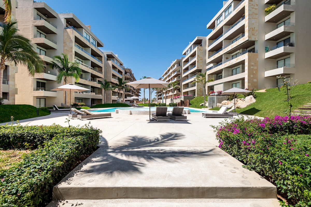 A spacious outdoor area is shown, featuring a central pool surrounded by lounge chairs and umbrellas. Lush greenery and blooming flowers line the pathway leading to the pool. The modern buildings of the complex create an inviting atmosphere under a clear blue sky.