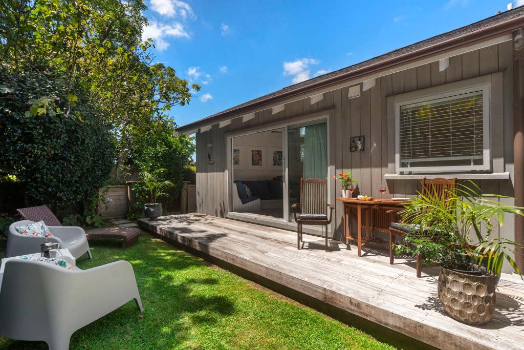 A private deck is visible, furnished with comfortable seating and a small dining table. Lush greenery surrounds the area, enhancing the tranquil ambiance. The living space behind features large glass doors, providing a seamless connection to the outdoor space.