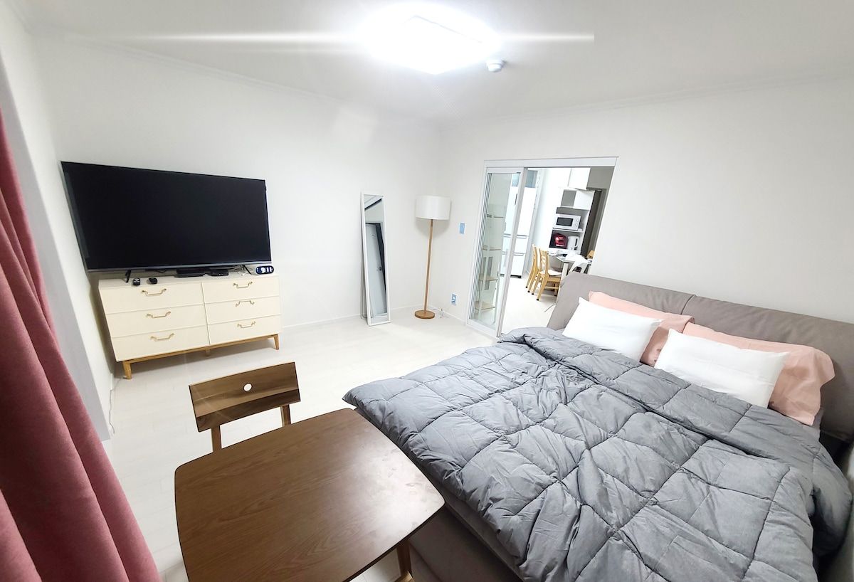 A spacious room features a comfortable bed covered with a gray quilt, complemented by pink pillows. A television is mounted on the wall across from the bed. A wooden table adds functionality, while a floor lamp provides additional lighting.