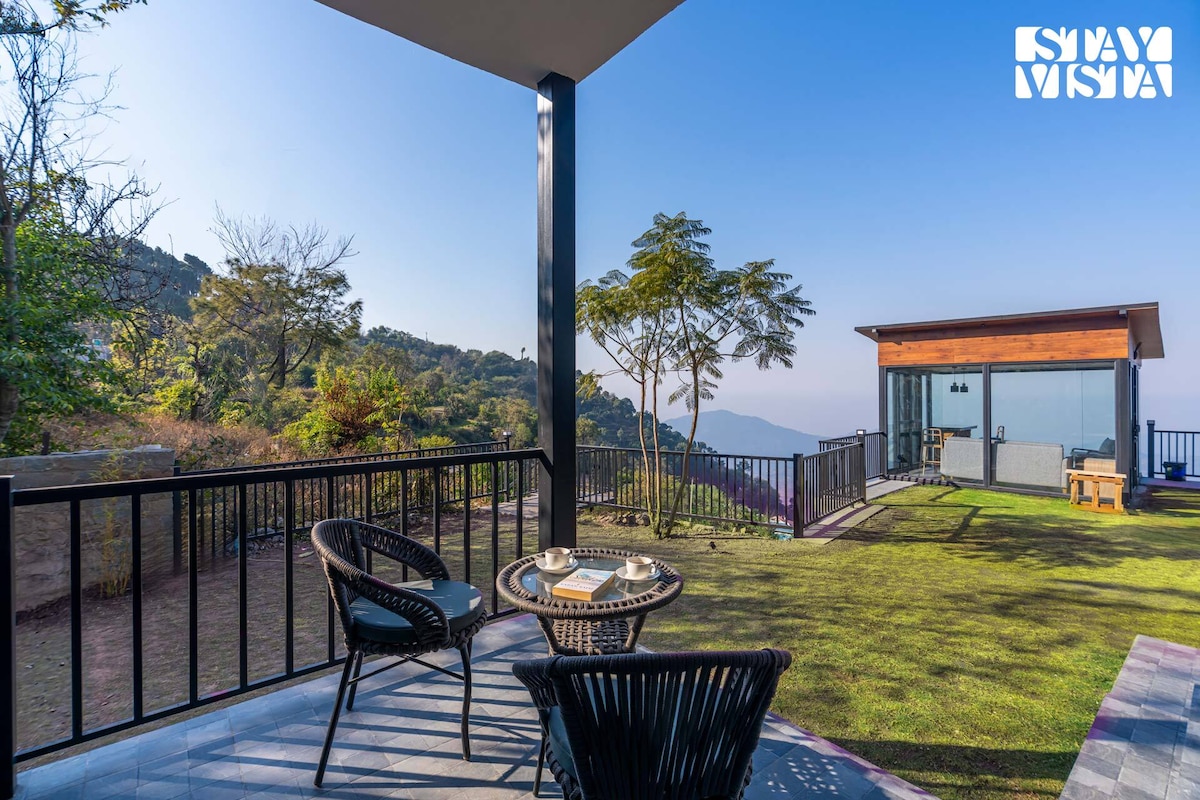 An outdoor seating area presents two black wicker chairs surrounding a small table set with two cups. The view features a vast green lawn bordered by trees and mountains in the distance under a clear blue sky, creating a serene atmosphere.