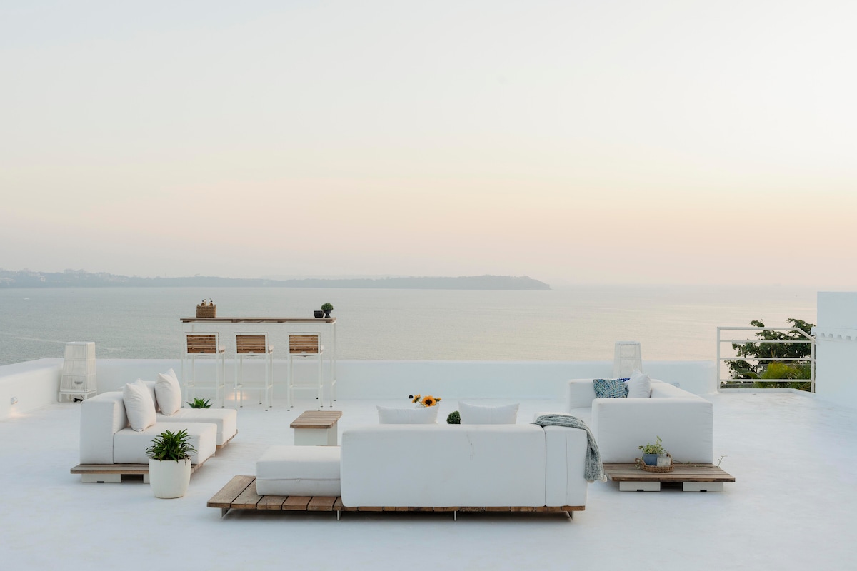 An expansive all-white deck features multiple lounge areas, inviting relaxation against a backdrop of the azure sea. Plush seating is arranged for comfort, with a sleek bar in the distance. The tranquil atmosphere is enhanced by the soft hues of the sunset sky.