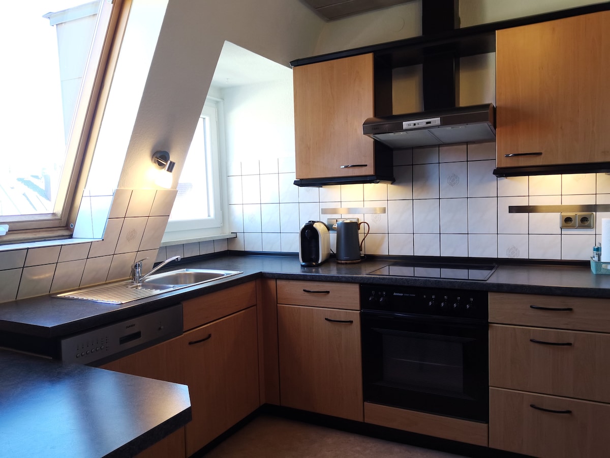 A modern kitchen features light wood cabinetry and dark countertops. Natural light enters through a large window beneath a slanted ceiling. Appliances include an electric stove, oven, and two small kitchen appliances, with ample counter space available for meal preparation.