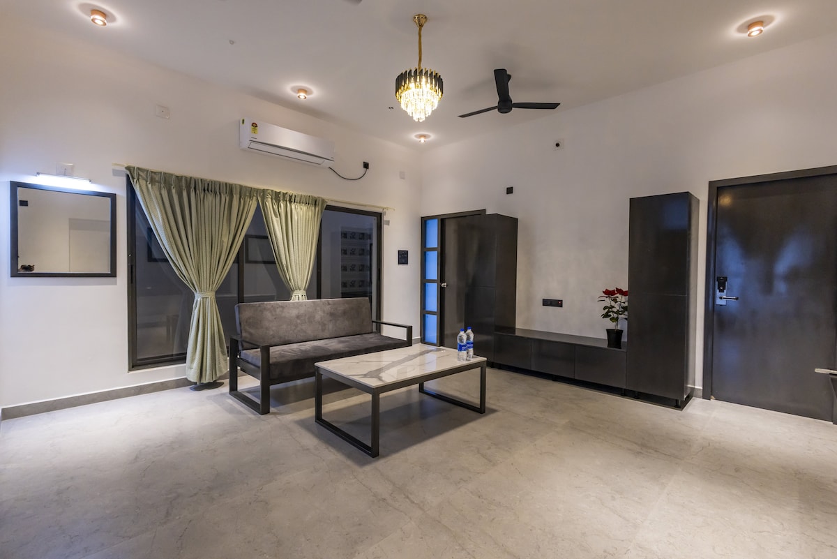 A spacious living area features a gray sofa and a coffee table, accentuated by natural light from large windows. Light-colored drapes frame the windows, and a ceiling fan provides airflow. A minimalist design is complemented by a sleek cabinet and a wall mirror.