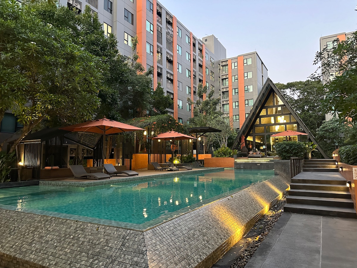 An outdoor area features a sparkling swimming pool surrounded by lush greenery. Multiple lounge chairs are positioned under orange umbrellas. A modern building with large glass windows rises in the background, reflecting soft evening light. Pathways lead to various landscaped sections of the facility.