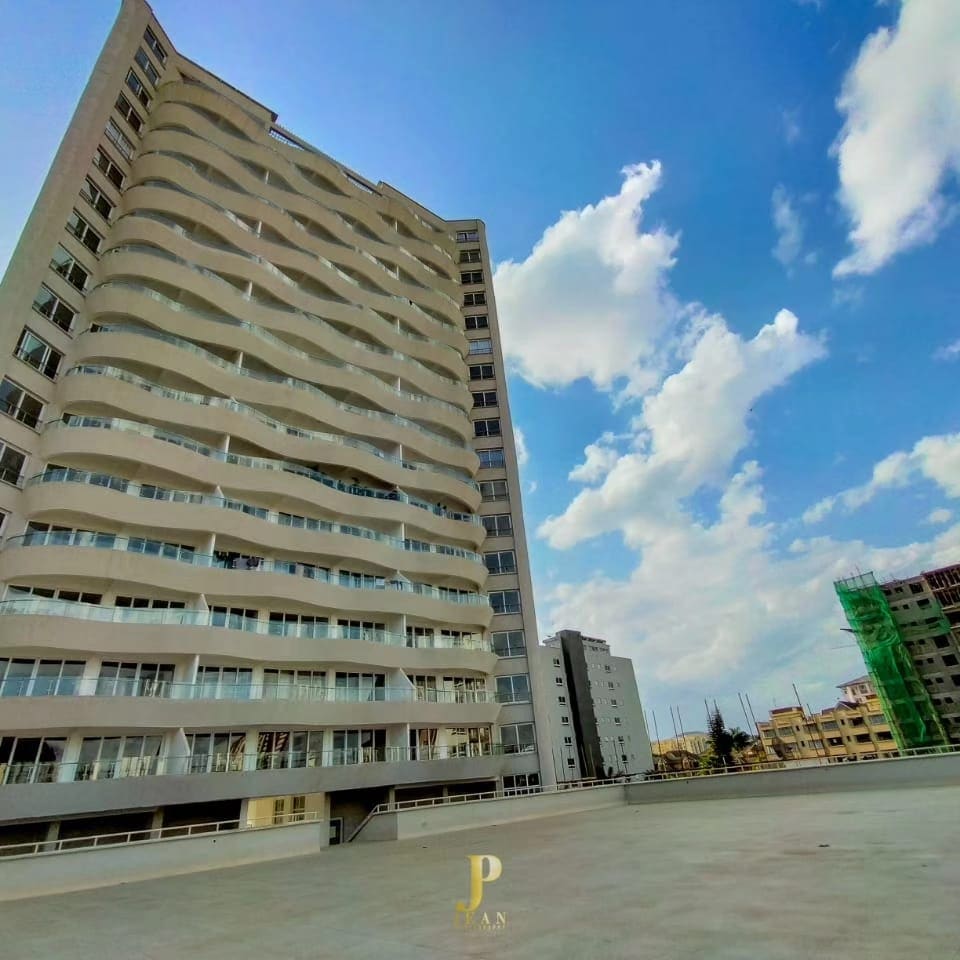 The modern building features a distinctive wavy façade, accentuated by expansive balconies. The structure rises impressively against a clear blue sky, while surrounding areas include construction activity and greenery, highlighting the development in the neighborhood.