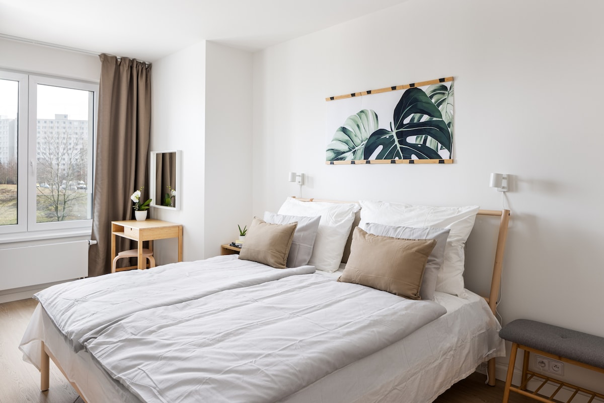 A spacious bedroom is presented with a large bed featuring neutral bedding and two decorative pillows. Natural light fills the space through a wide window, complemented by soft curtains. A minimalist desk and mirror add functionality, while a tropical print artwork enhances the decor.