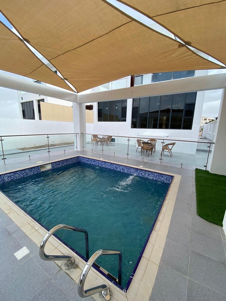 A modern swimming pool is positioned under a shaded area, surrounded by sleek tiles and glass railings. Nearby, a dining space with multiple wicker chairs is visible, allowing for relaxation and social gatherings. Large windows provide natural light and views of the outdoors.