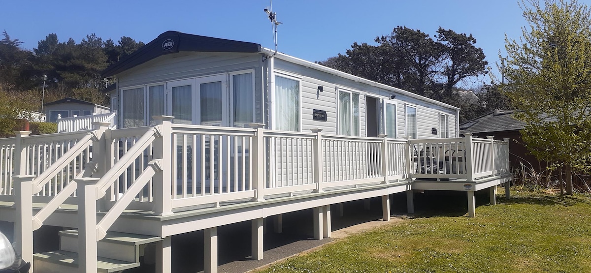 A luxury caravan is presented on a raised deck, surrounded by well-maintained greenery. The wrap-around decking features sturdy white railings, providing a spacious area for outdoor relaxation. The structure showcases large windows, allowing for ample natural light and views of the surrounding landscape.