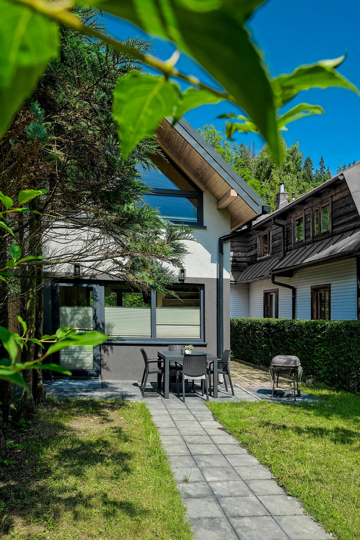 The exterior of the house is visible, featuring large windows that allow natural light to enter. A paved path leads to a patio area with outdoor furniture, and a barbecue grill is positioned nearby. Lush greenery surrounds the space, enhancing the tranquil setting.