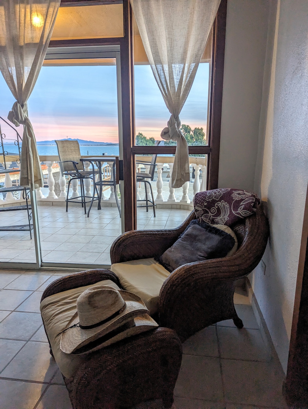 A comfortable wicker chair is positioned next to a small footrest, with a hat resting on it. Sheer curtains frame a large glass door, revealing a balcony with outdoor seating and a view of the sea at sunset.