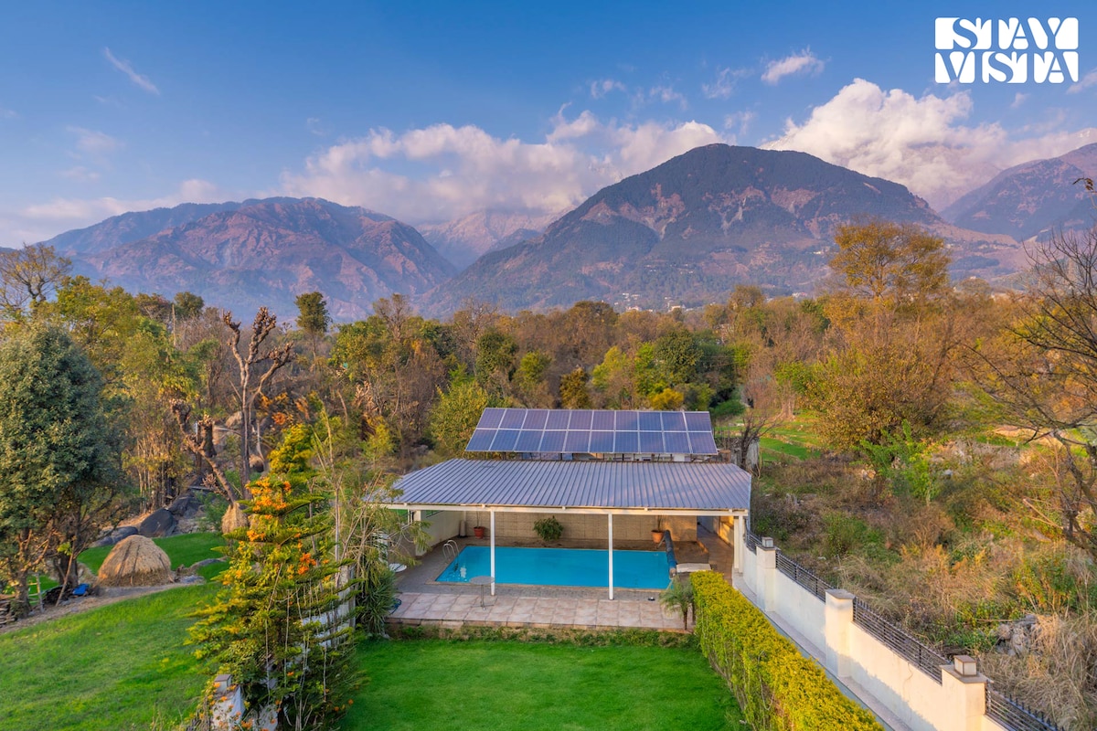 The outdoor area features a swimming pool nestled among lush greenery, framed by scenic mountains in the background. Solar panels are installed on the roof, reflecting a commitment to sustainability. The open space invites relaxation and enjoyment of the serene natural surroundings.