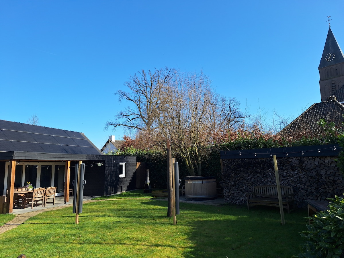 A spacious garden area features a well-maintained lawn, with seating arrangements made from wood. A hot tub is visible, surrounded by verdant greenery and a wooden fence. In the background, a traditional church tower is partially seen against a clear blue sky.