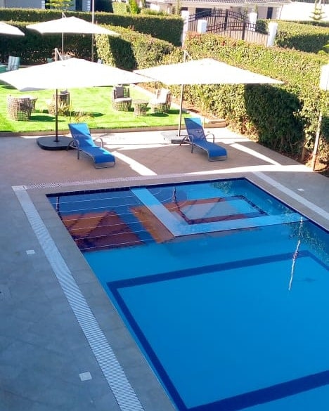 A swimming pool is featured surrounded by a well-manicured lawn. Comfortable lounge chairs are positioned under large umbrellas, providing shaded seating areas. The pool is designed with a geometric pattern, and steps are visible leading into the water.