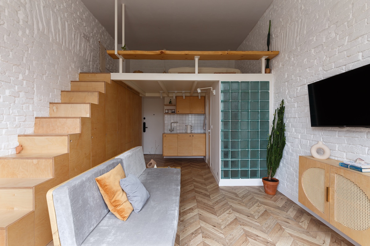 A spacious living area features a light-colored sofa with accent pillows, complemented by natural wood elements. A staircase leads to a lofted sleeping area above. A glass block wall provides natural light, while a potted plant adds a touch of greenery to the corner.