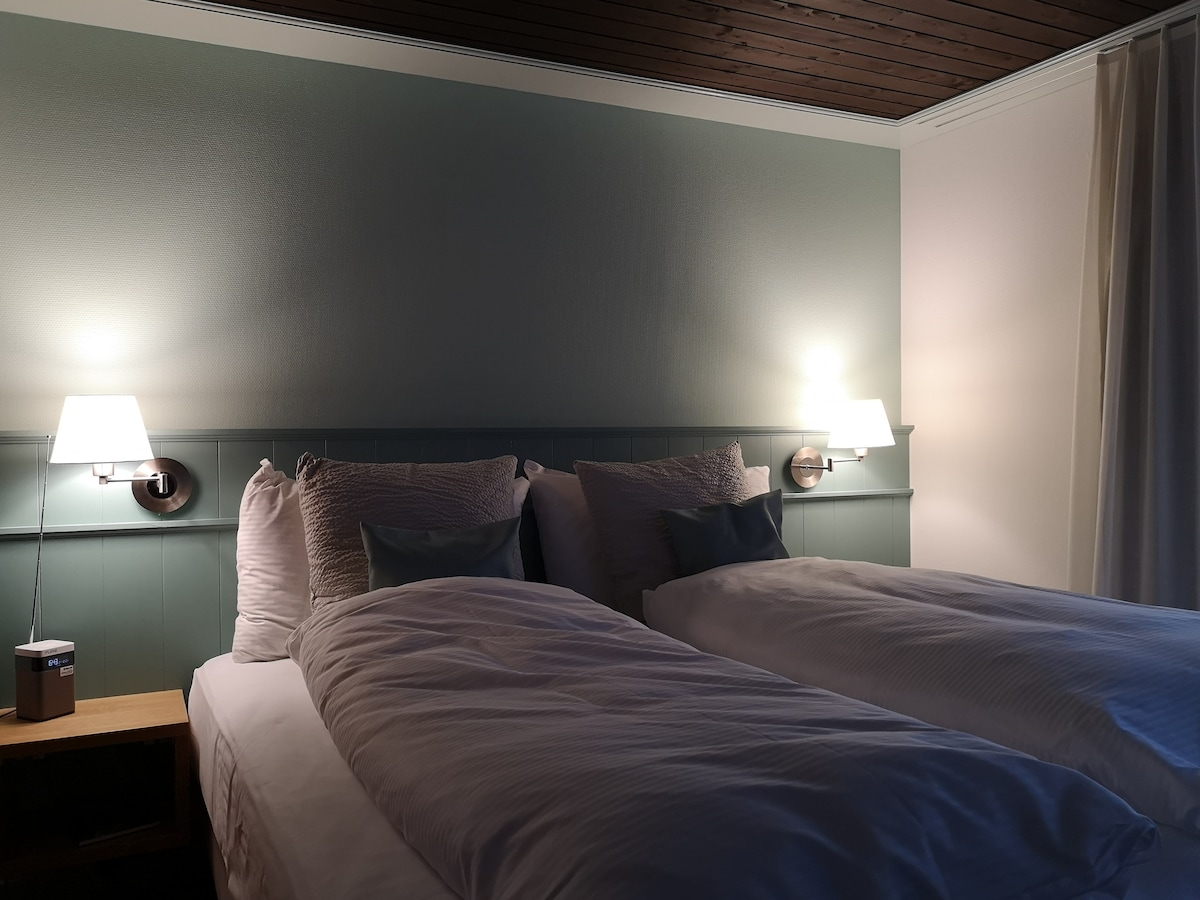 A serene sleeping area is presented, featuring two spacious beds neatly made with white linens. Each bed is accompanied by a bedside lamp. Soft green walls add a calming touch, while dark wooden beams contrast with the light colors, contributing to a restful ambiance.