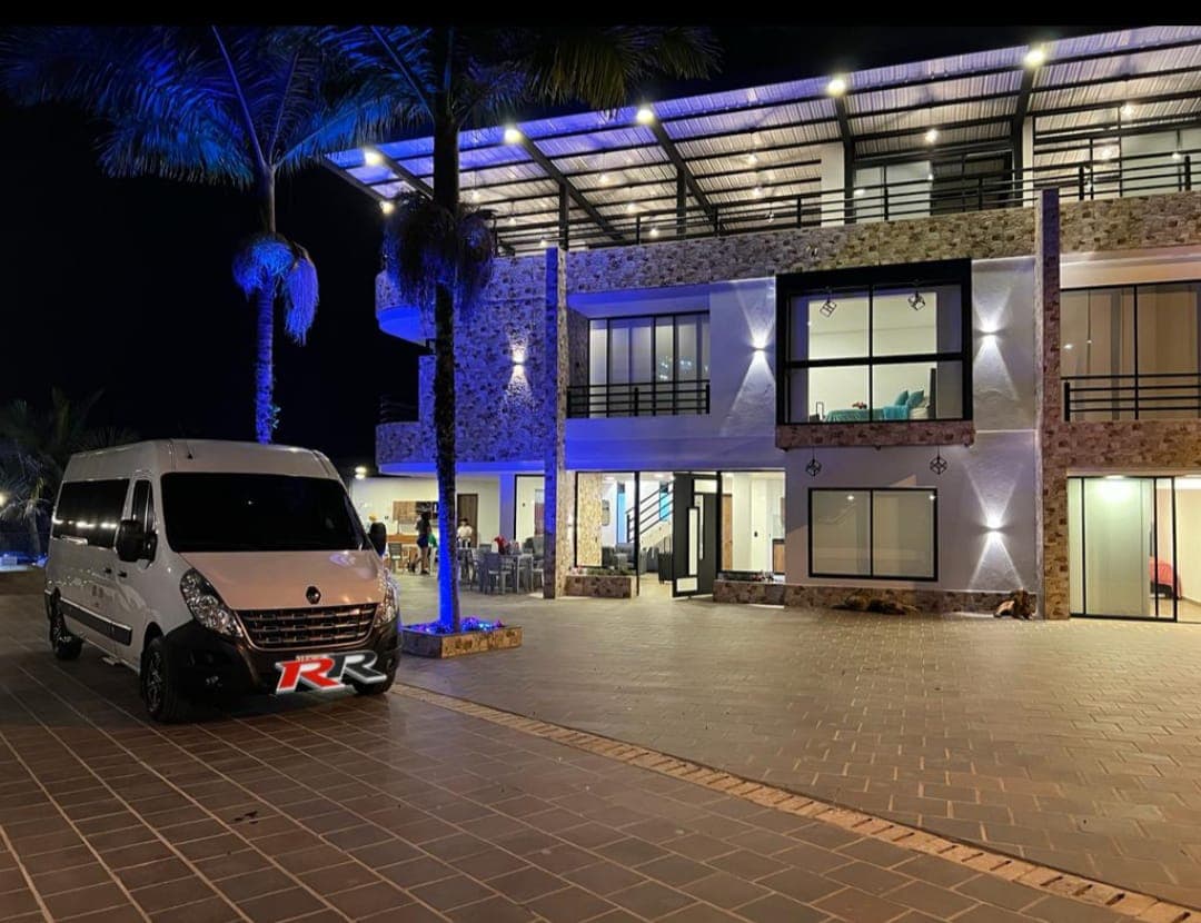 A well-lit exterior showcases a modern multi-story building with a stone facade. Palm trees frame the entrance, where a white vehicle is parked on the patterned floor. The spacious outdoor area features ample seating arrangements visible through large windows.