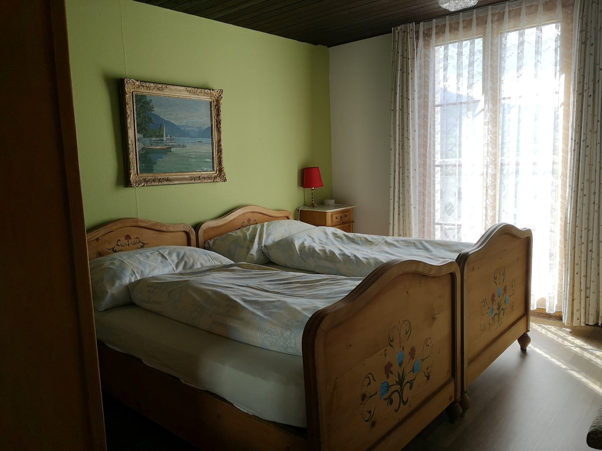 A sleeping area features two wooden beds with intricate floral carvings, dressed in light bedding. Soft light filters through sheer curtains, illuminating the space. A small red lamp sits on a bedside table, and a landscape painting adorns the green wall.