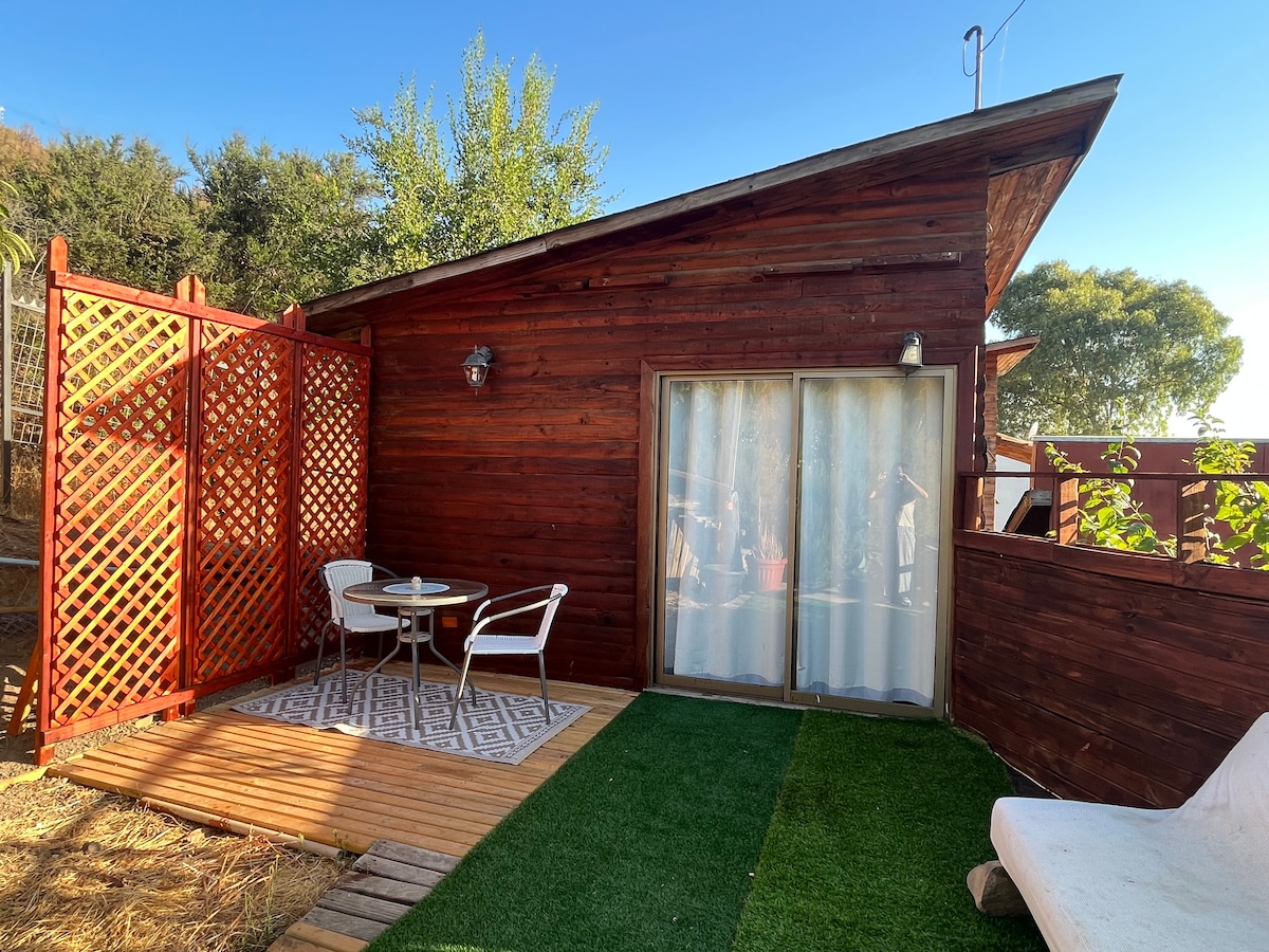 An outdoor terrace features a small table and two chairs, surrounded by wooden fencing. A large glass door provides access to the interior, while artificial grass adds a touch of greenery alongside wooden decking. Sunlight brightens the space, creating a welcoming entrance.