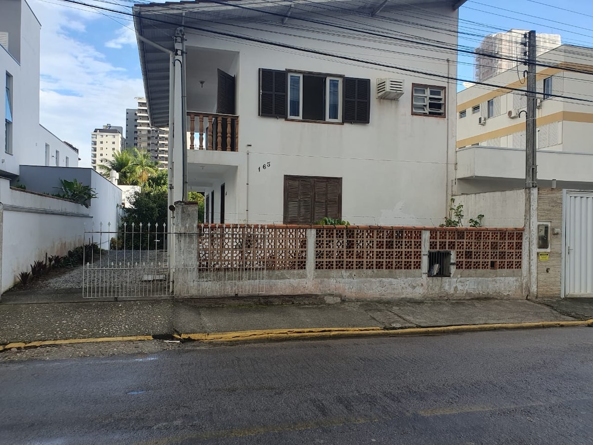 The exterior of a two-story house features a white facade with dark shutters and a balcony. A gated entrance leads to a garden area with gravel, while a sidewalk adjoins the street. Nearby buildings provide a glimpse of the local architecture.