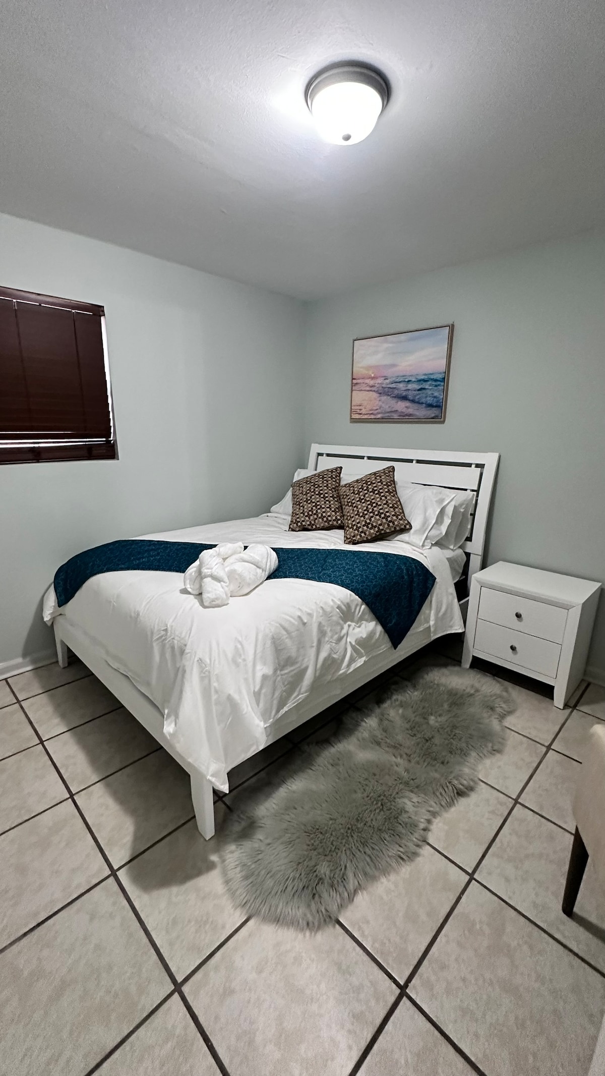 A bright bedroom features a spacious bed with white linens, accented by decorative pillows. A bedside table with a lamp stands next to the bed. Natural light enters through a window with brown blinds. A soft rug adds texture to the tiled floor, enhancing comfort.