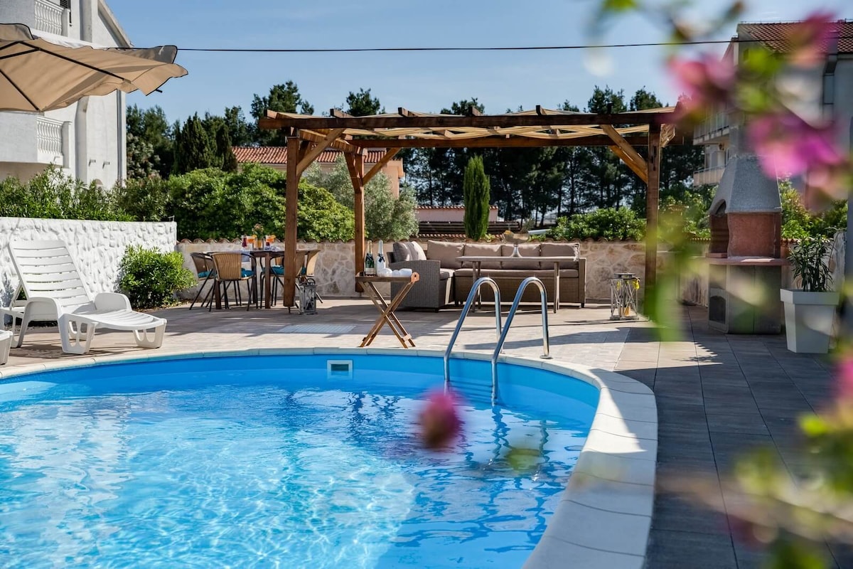 A private pool is surrounded by a spacious patio featuring lounge chairs and a shaded seating area under a wooden pergola. A nearby dining table accommodates several guests, providing an inviting space for outdoor leisure and relaxation.