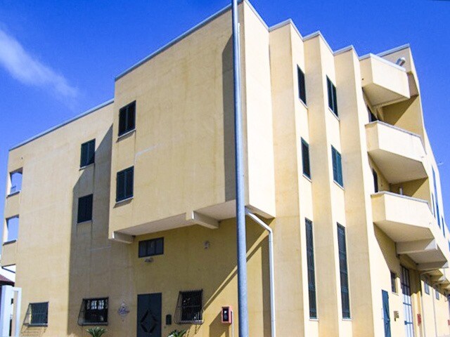 The exterior of a three-story building is depicted, featuring a yellow facade with multiple large windows. Balconies are seen on the upper levels, and the structure is set against a clear blue sky, indicating a bright and open environment.