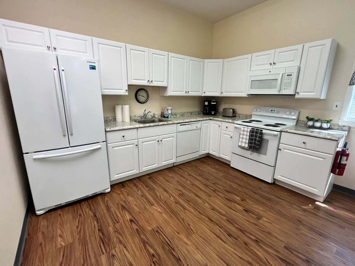 A bright kitchen features white cabinets and a granite countertop. A refrigerator, dishwasher, and stove are situated along the walls, while a clock adds a functional touch. The wood-style flooring complements the space, enhancing the overall contemporary feel.
